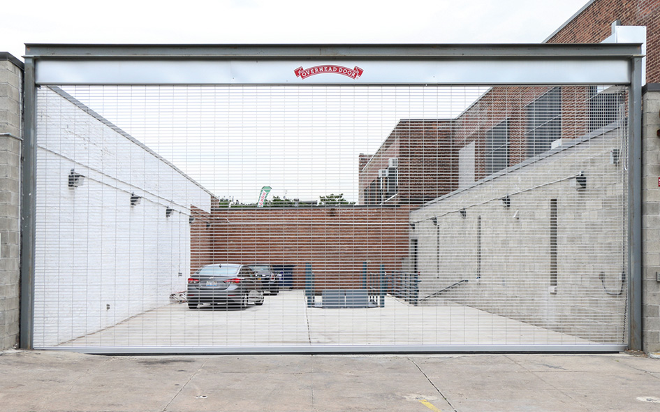 A silver Model 670 security grille is lowered across the entrance to a parking lot, with two cars and a brick building visible behind it. The upward coiling gate features a red and white sign at the top center.