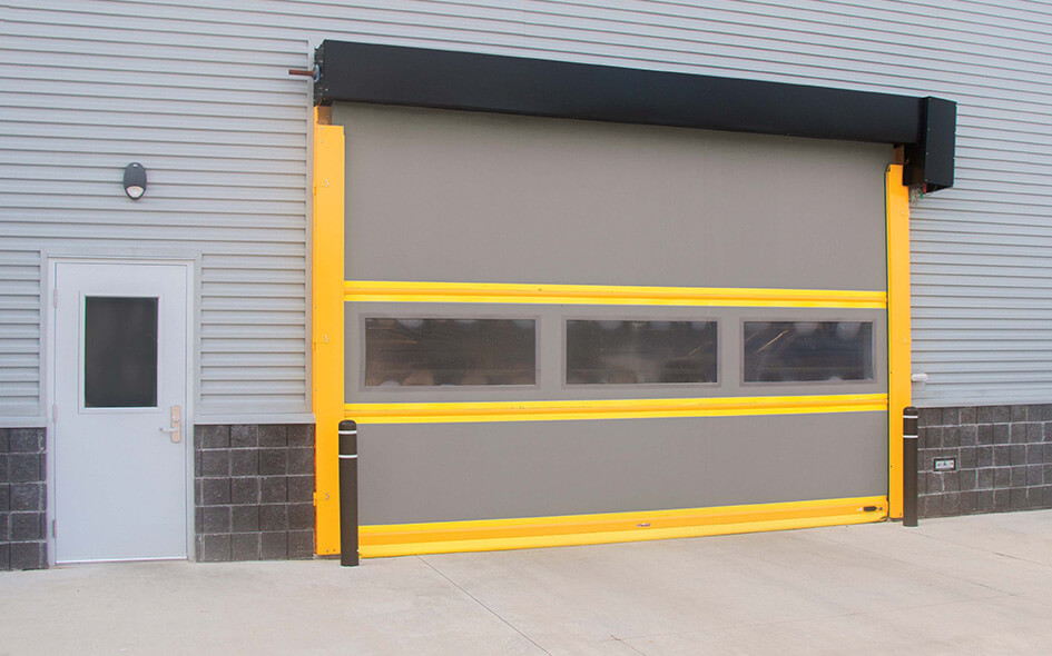 A large High Speed Door with yellow safety edges and transparent panels is installed on a gray building exterior, next to a standard white entry door.