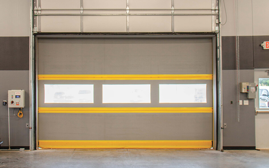 A large high speed exterior door with yellow horizontal safety stripes and three rectangular windows, inside a warehouse with gray walls and visible control panels on the left.