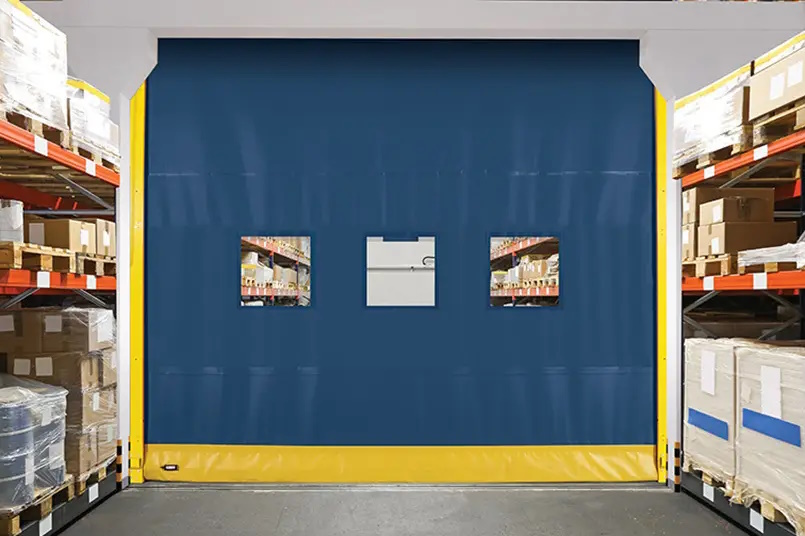 A blue Flexible Bottom High Speed Door with two small windows stands in a warehouse, surrounded by shelves stacked with wrapped pallets and boxes.
