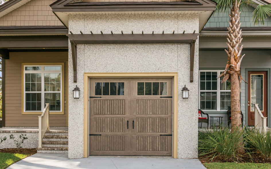 A single-car garage with an Impression Fiberglass Collection wooden-look door, decorative lanterns on each side, and a pergola-style overhang attached to a house with steps, windows, and a palm tree nearby.