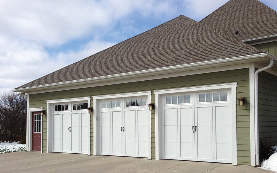A three-car garage with white paneled doors and small windows at the top, attached to a house with light green siding and a shingled roof, under a partly cloudy sky.