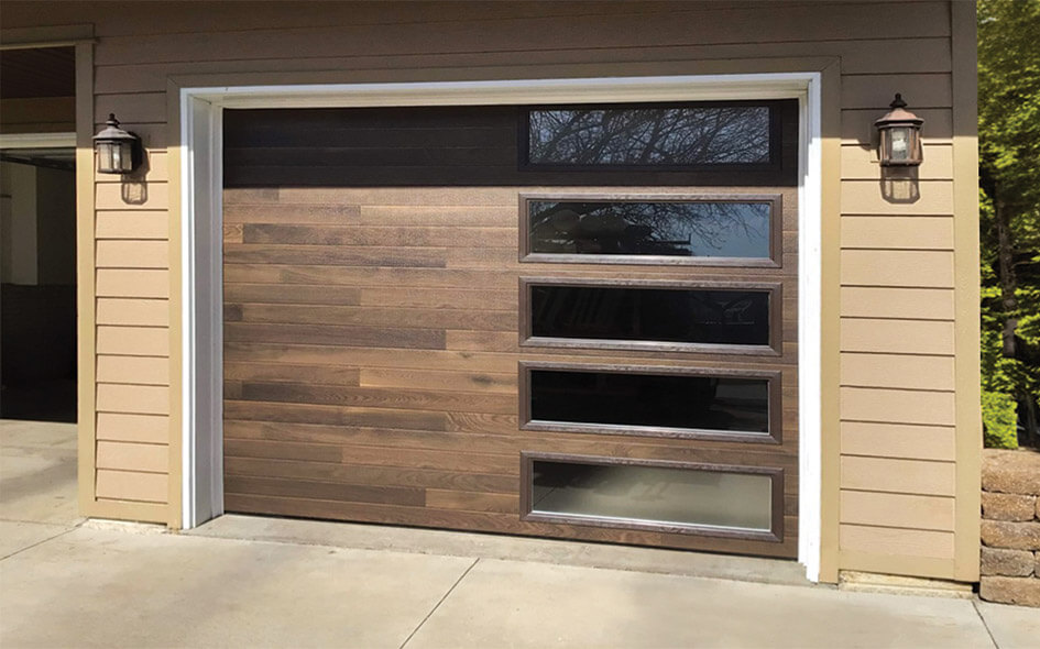 A brown wooden Thermacore Collection garage door with four horizontal rectangular glass windows on the right side, set in a beige house exterior with two wall-mounted lantern lights on either side of the door.