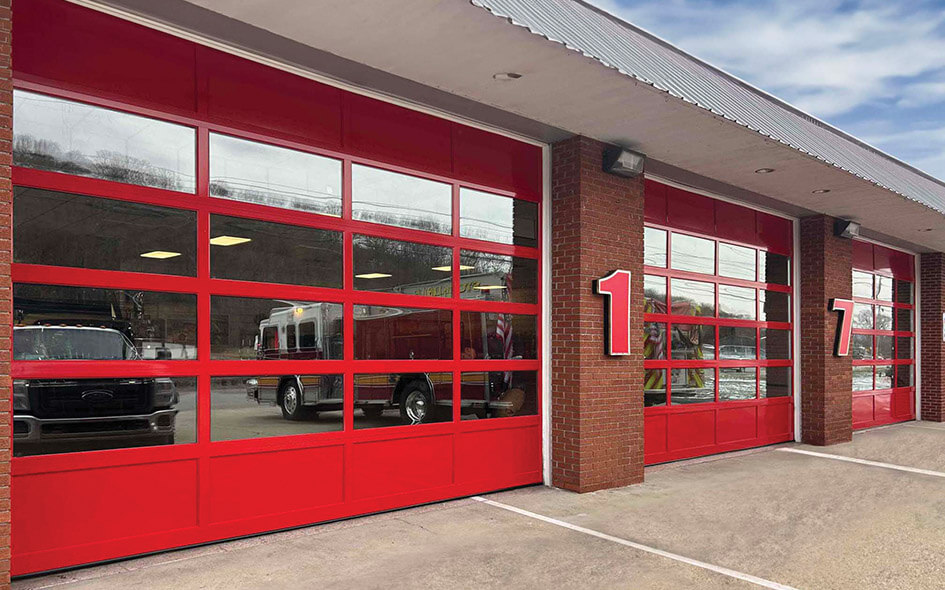Three large red garage doors with glass panels are set in a brick fire station. Fire trucks are visible inside. The number 1 is mounted between the first and second doors. The sky is partly cloudy.