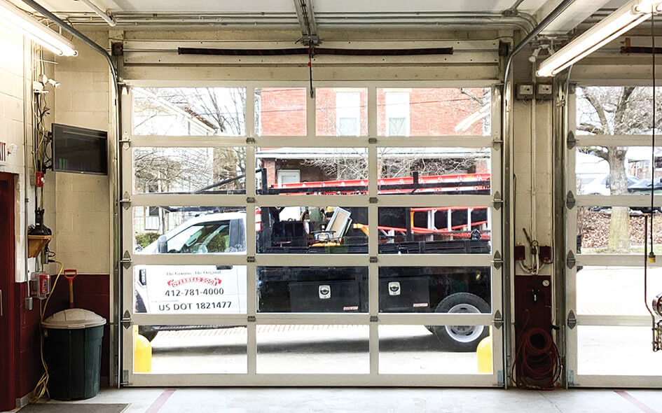A black utility truck with red ladders on top is parked outside a glass-paneled garage door. The truck has a sign with a phone number and USDOT number. Trees and a brick building are visible in the background.