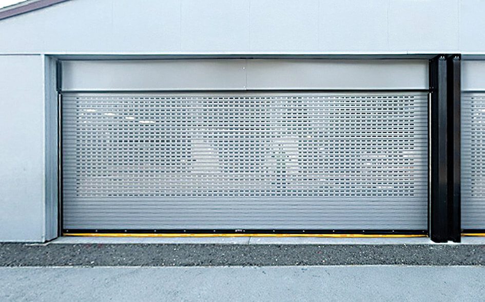 A silver perforated Rolling Steel Door 610 is partially closed on a modern industrial building, with a paved area in front and a smooth, light-colored exterior wall—showcasing the versatility of service doors.