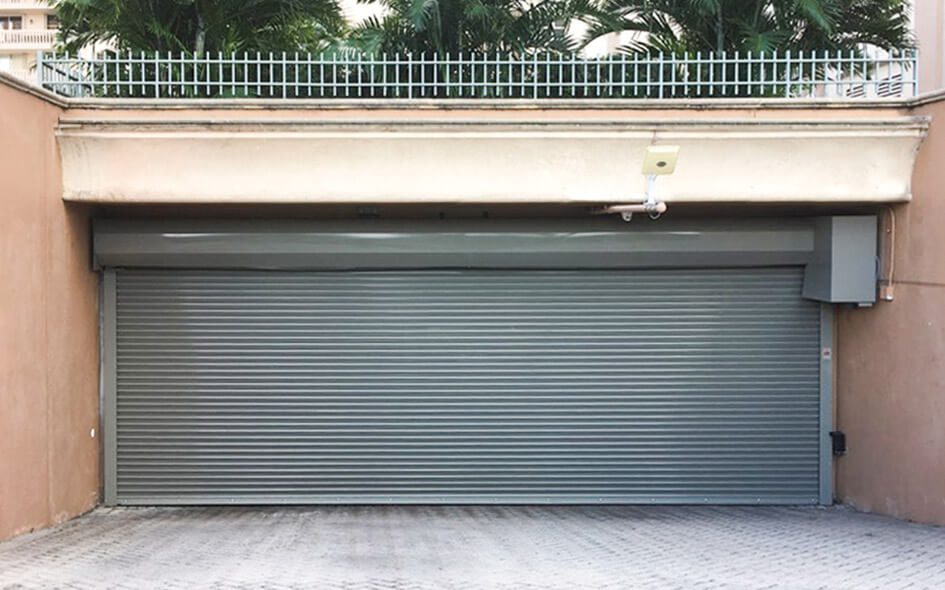 A closed gray metal roll-up garage door, likely a 610 Rolling Steel Door, set in a peach-colored wall with a security camera above and palm trees visible behind a metal fence.