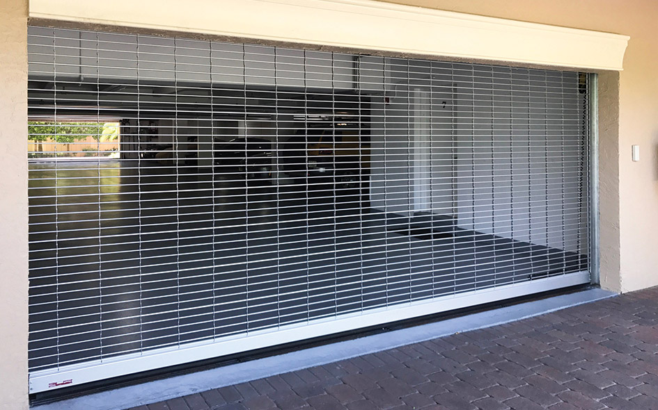 A silver metal model 670 security grille gate with an upward coiling design covers the entrance of a garage with a brick driveway, allowing visibility into the empty, well-lit interior space behind it.