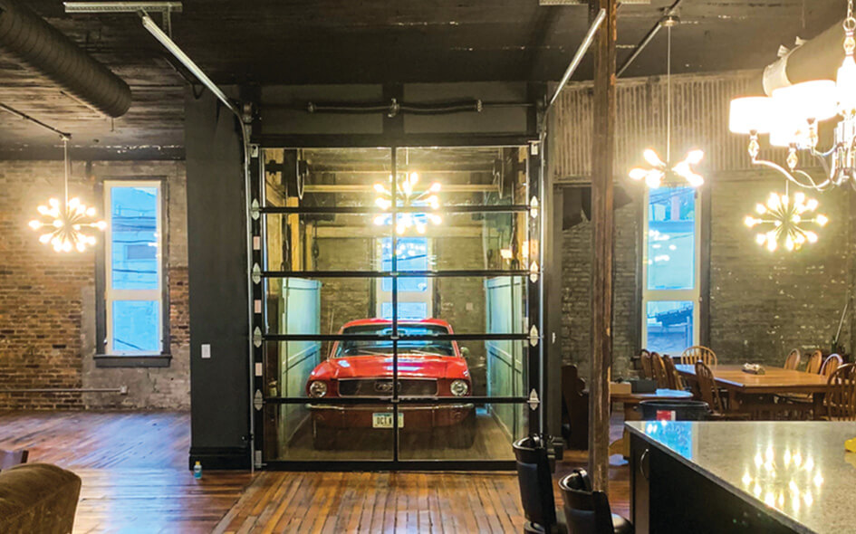 A red vintage car is showcased behind a modern aluminum and glass garage door inside a stylish loft apartment with exposed brick walls, wooden floors, modern lighting fixtures, and a large dining table.