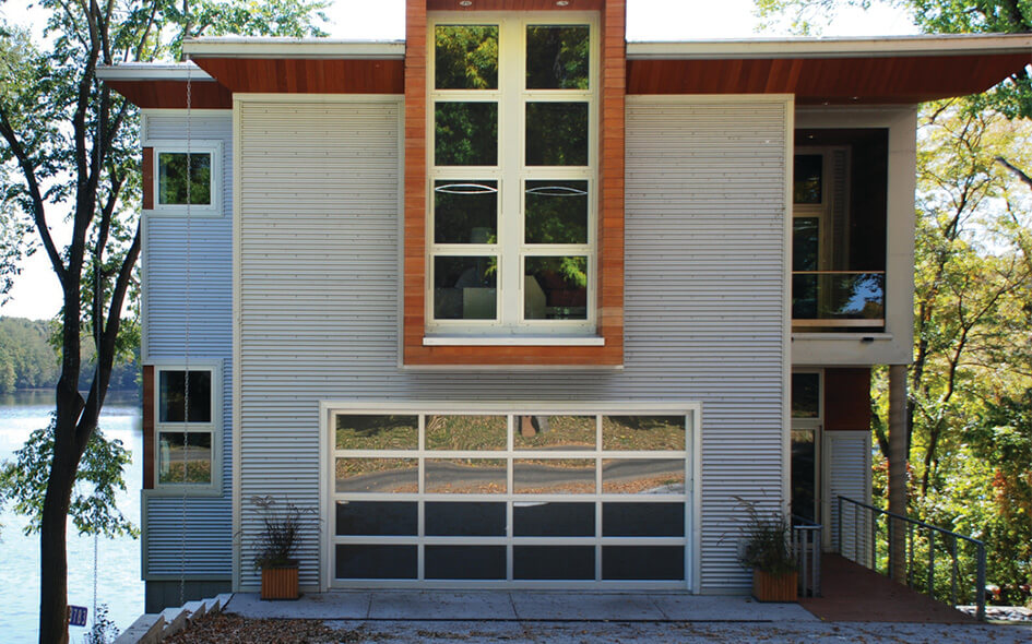 Modern house with large windows and a prominent modern aluminum and glass garage door, surrounded by trees and overlooking a lake, with sunlight filtering through the foliage.