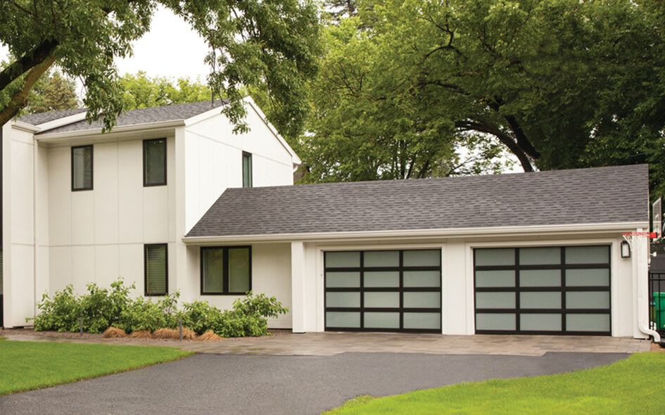 Modern two-story white house with dark trim and a large attached garage featuring three modern aluminum frosted glass doors. The home is surrounded by green grass, shrubs, and tall trees.