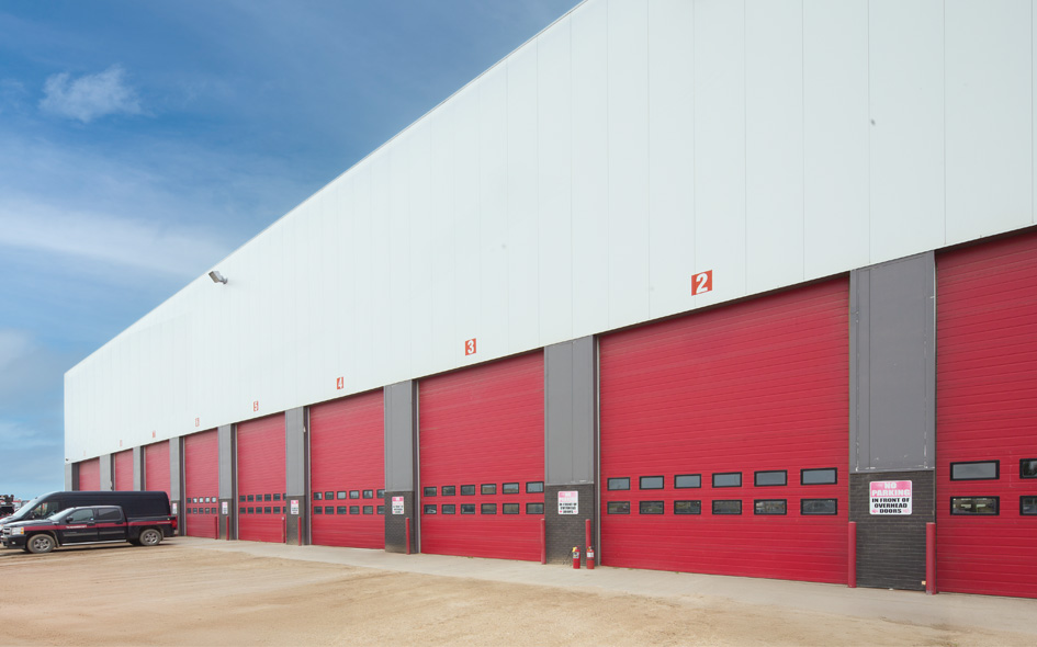A row of large red Thermacore sectional steel doors, each labeled with numbers like 592, lines a white building. A black van is parked to the left beneath a clear sky.