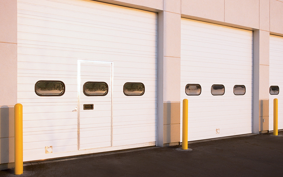 Three large white Sectional Steel Doors with narrow rectangular windows, set in a beige building. Yellow safety bollards stand in front of each door. The evenly lit scene features the durable Thermacore 592 design, with no visible people or vehicles.