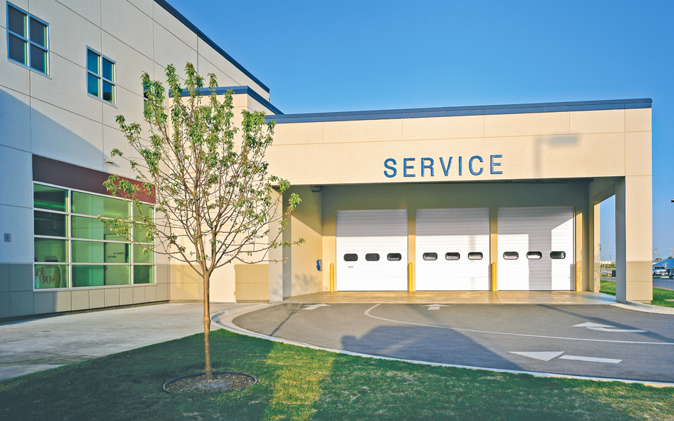 Modern building with three white Thermacore sectional steel doors labeled SERVICE, a small tree in the foreground, and clear blue sky above. Arrows are painted on the driveway leading to the service area.