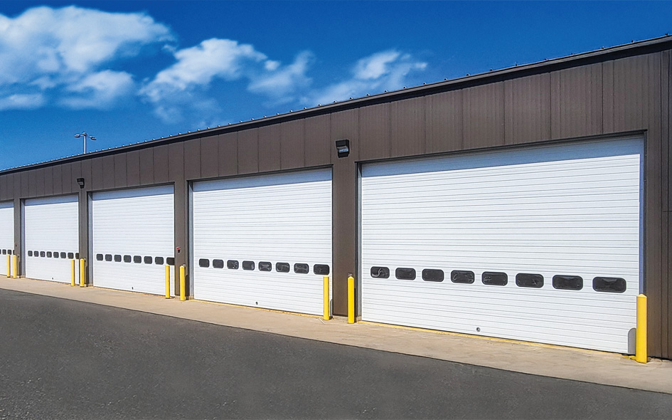 A row of large white Thermacore sectional steel doors on a brown metal building, with yellow bollards in front and a clear blue sky with a few clouds above.