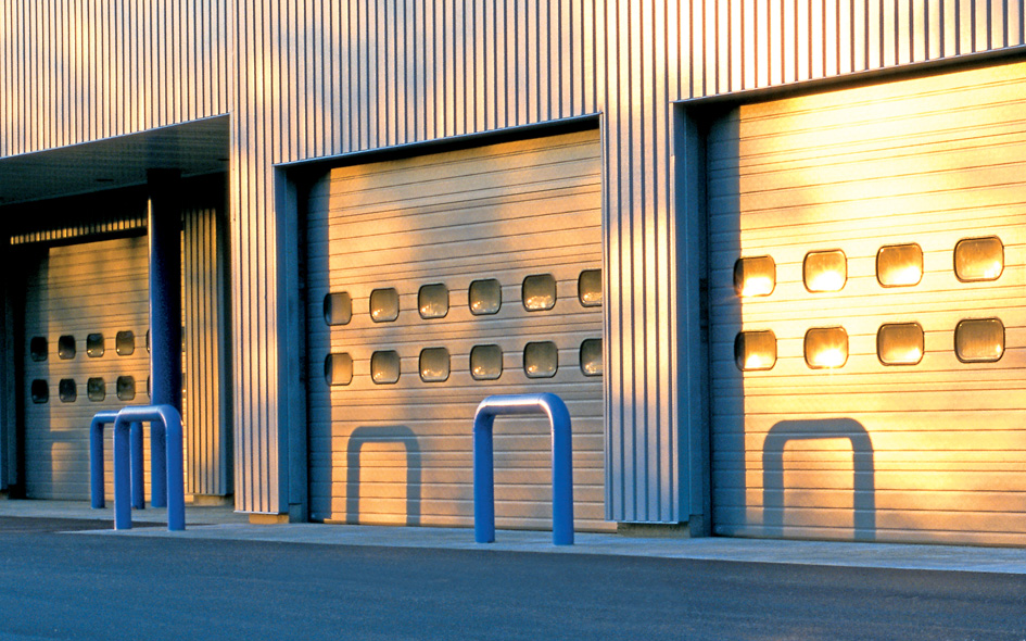 Three Thermacore 592 sectional steel doors with small rectangular windows are closed on a metal building. Blue bollards stand in front of each door, all bathed in warm sunlight.