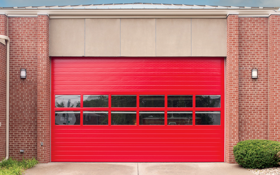 A large, bright red sectional steel door with rectangular windows along the middle, set in a brick building with two wall lights and some bushes on the right side. This Thermacore model 592 offers both durability and style.