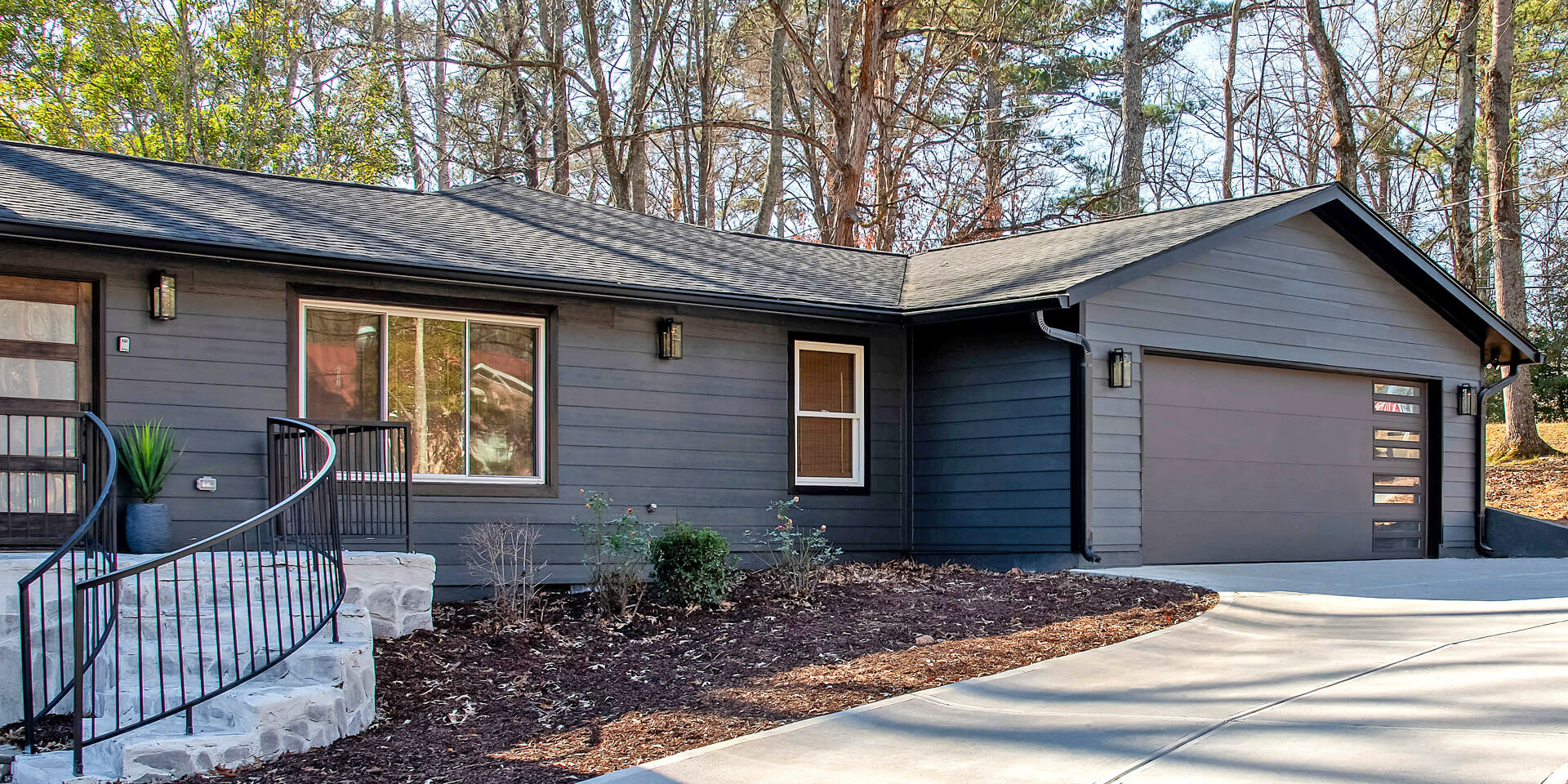 Single-story modern house with dark gray siding, a large front window, Thermacore Collection insulated garage doors, curved stone steps with metal railing, and a driveway, surrounded by trees and minimal landscaping for an energy efficient home.
