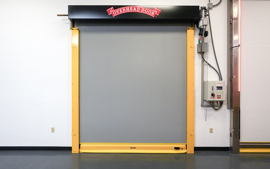 A yellow high speed door with a gray panel is closed, set in a white wall. Above the door is a black sign with a red label reading OVERHEAD DOOR. Electrical panels and exposed wiring are mounted beside this 991-series entry.