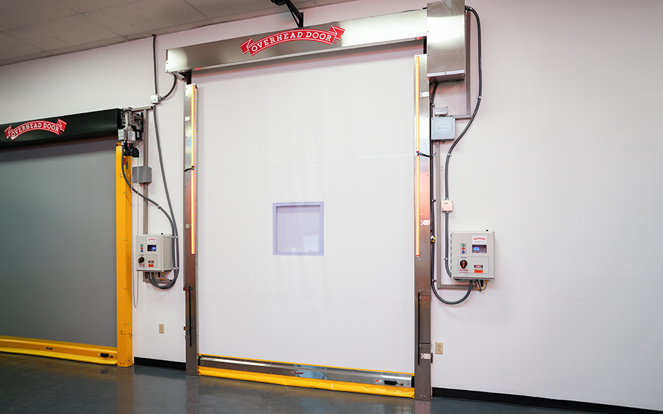 A large, white High Speed Door with a small square window and metal frame, labeled Overhead Door, is installed in a facility. Control panels sit on either side, along with a yellow safety barrier at the base. Model: 991.