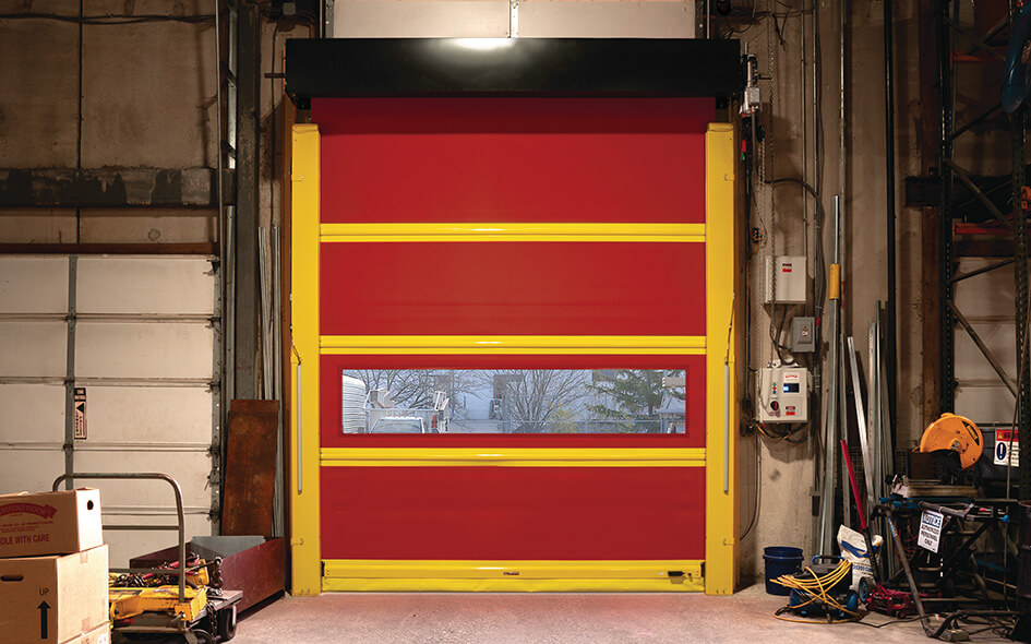 A bright red High Speed Door with yellow borders, model 993, is closed inside a warehouse. The Exterior Fabric Door features a clear panel revealing trees and a house outside. Equipment and boxes are scattered around the space.