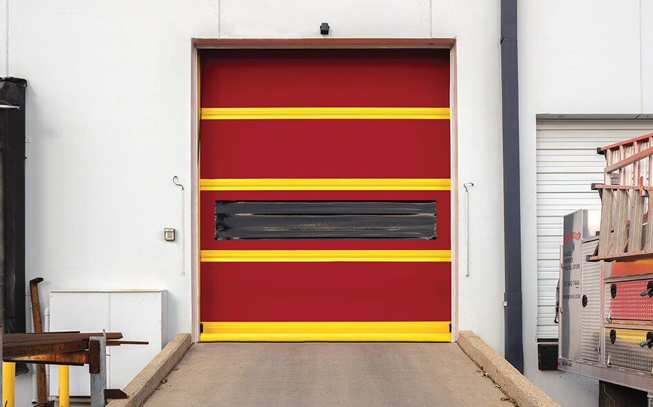 A bright red 993 High Speed Door with yellow horizontal stripes and a dark rectangular window, set in a white building. The concrete ramp leads to the door. Nearby are metal structures and a parked utility vehicle.