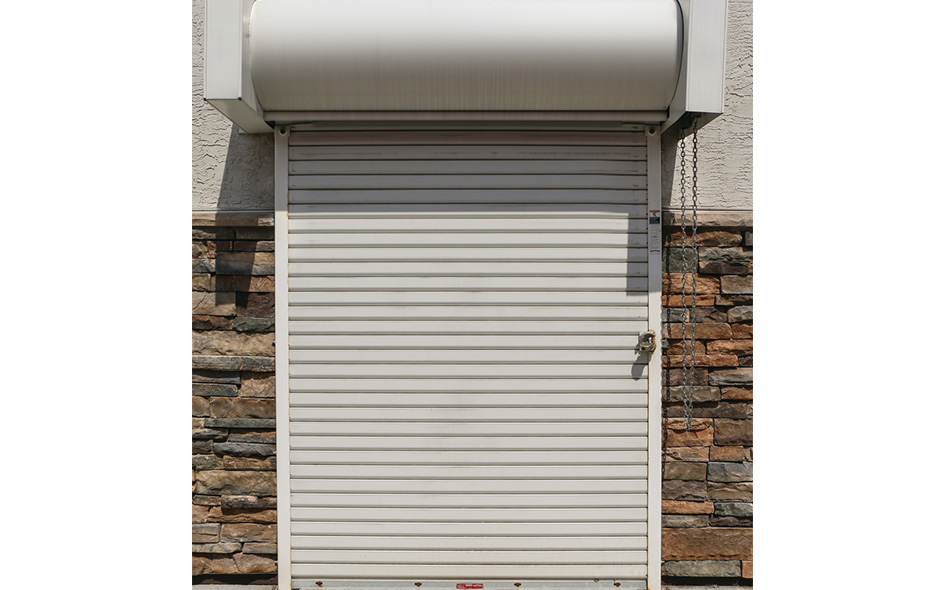A closed white Rolling Steel Door with a lock is set in a wall featuring beige stucco on top and a lower section of brown and gray stacked stone. A metal chain hangs on the right side.
