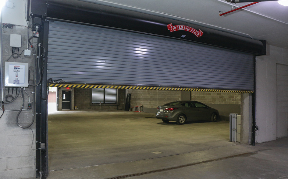 A partially open gray Model 611 overhead garage door reveals a parked gray car inside a concrete, indoor parking garage with cinder block walls and fluorescent lighting.
