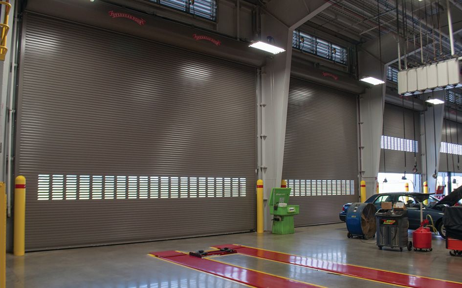 Large industrial garage with three closed Model 625 rolling steel service doors, a green machine, car lift, vehicle parts, and tools scattered on the floor inside a well-lit auto shop.