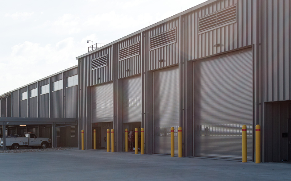 A large industrial building with multiple closed Model 625 Rolling Steel Doors, vertical siding, and yellow safety bollards in front. A small utility vehicle is parked on the left side near the building.