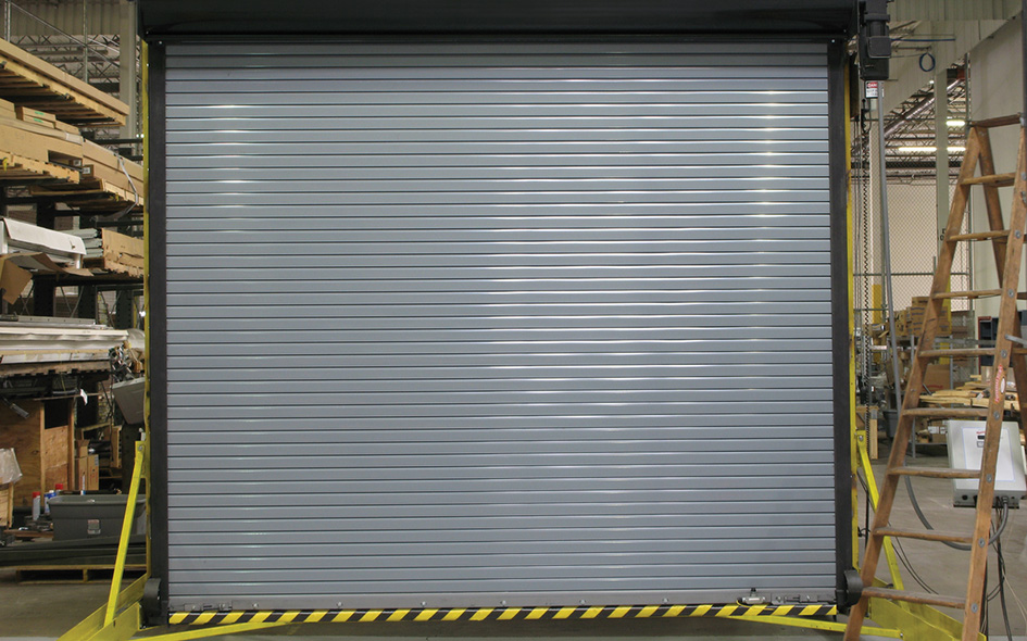 A large, silver Rolling Steel Service Door, Model 626, is closed inside an industrial warehouse. Yellow safety rails and black-and-yellow caution tape are visible at the base. Shelving and equipment surround the area.