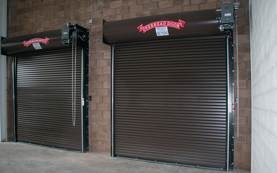 Two brown Rolling Steel Service Doors, Model 627, are installed side by side in a brick-walled building. Both doors are closed, each featuring an overhead motor unit labeled Overhead Door.