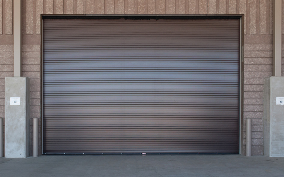 A large closed brown Model 620S EverServe industrial roll-up garage door is set in a light brown brick wall, with concrete pillars on either side and signs posted on both pillars.