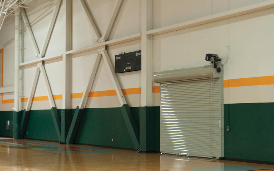 A gymnasium with green and orange walls, a scoreboard mounted high up, and a closed Rolling Steel Service Door. The polished wooden floor reflects light from above.