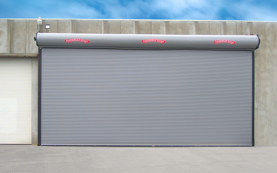 A large gray Rolling Steel Door is closed on a concrete warehouse building. The sky is partly cloudy above, and a smaller white Service Door, Model 620, is visible on the left side.