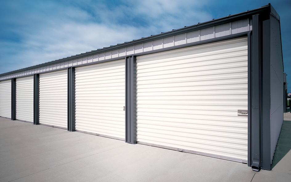 A row of closed white Model 790CW rolling sheet doors on storage units in a metal building, with a concrete driveway and a partly cloudy sky above.