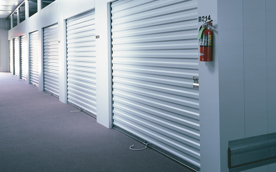 A row of closed white Model 770SS Rolling Sheet Doors lines the carpeted hallway, each labeled with a number. A fire extinguisher is mounted on the wall near door B214.