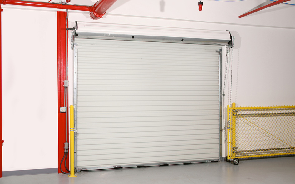 A closed white Model 780CD rolling sheet door is set in a white wall with red pipes above and along the side. A yellow safety barrier and chain-link fence are positioned to the right of the door.