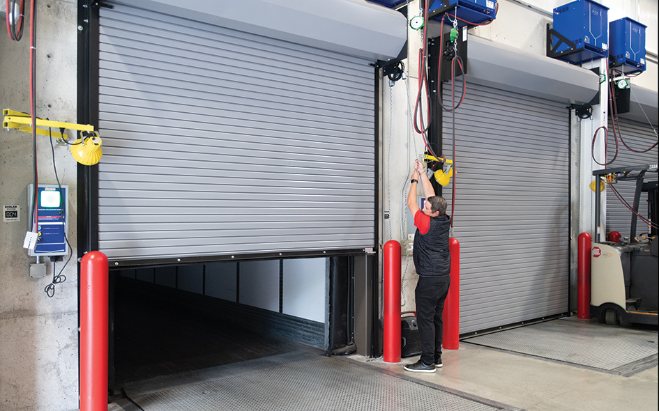 A person operates a pull chain to open a large metal Model 625 rolling steel door in a warehouse loading dock area. Red protective bollards and control panels are visible near the service doors.