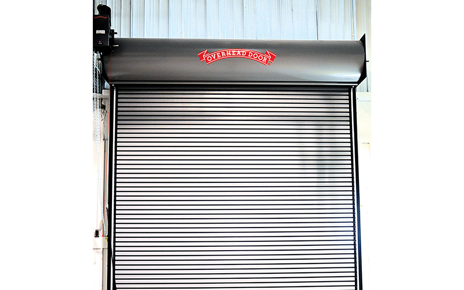 A metallic Model 627 industrial roll-up overhead door is fully closed, with a red sign at the top that reads Overhead Door. The background features a light-colored, corrugated wall.