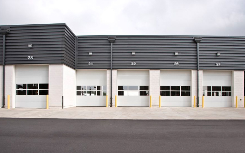 A row of five large, closed Model 418 sectional steel doors numbered 23 to 27 on a modern industrial building with a gray metal facade, white walls, and yellow safety bollards in front.