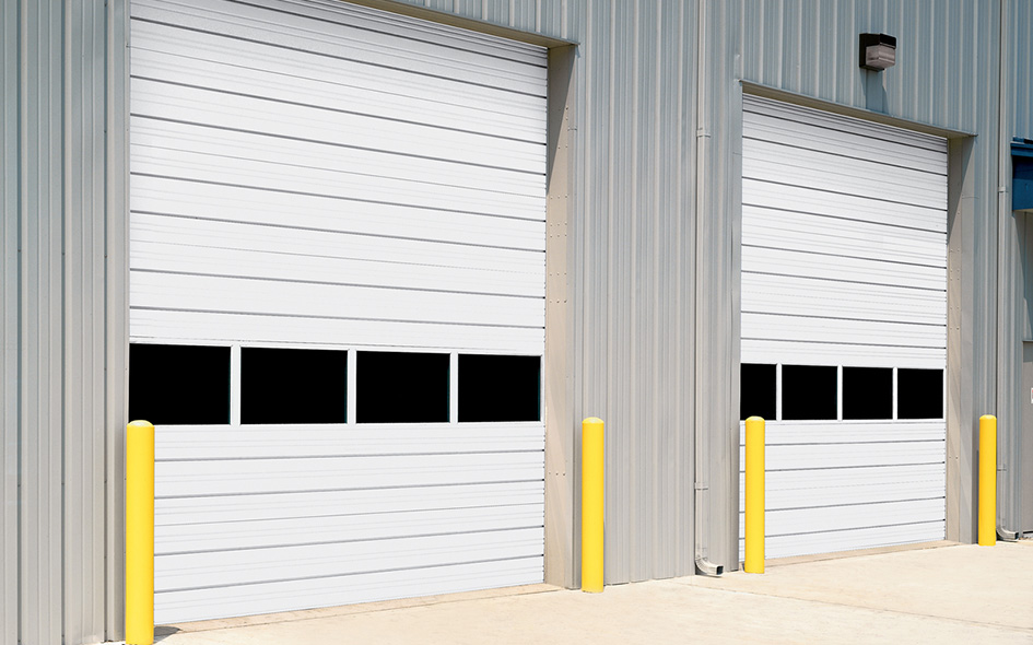 Two large white Model 432 sectional steel doors with horizontal panels are set on a metal building. The left steel door features four windows, while yellow safety bollards stand before both doors on the concrete surface.