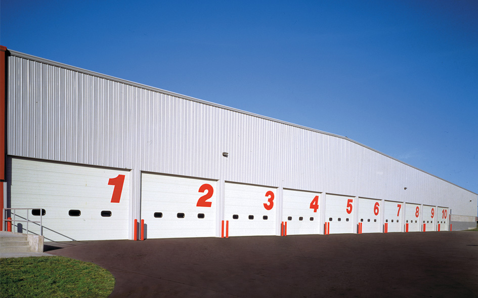 A row of ten large white Model 422 sectional steel doors, each labeled with a red number from 1 to 10, lines the white industrial building against a clear blue sky.