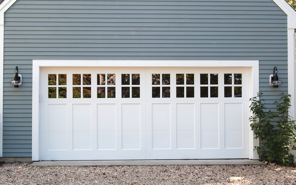 A white Signature Carriage double garage door with rectangular windows at the top, set in a light blue house with horizontal siding. Two wall-mounted lantern lights flank the door, and some greenery lines the right edge. Gravel covers the ground.