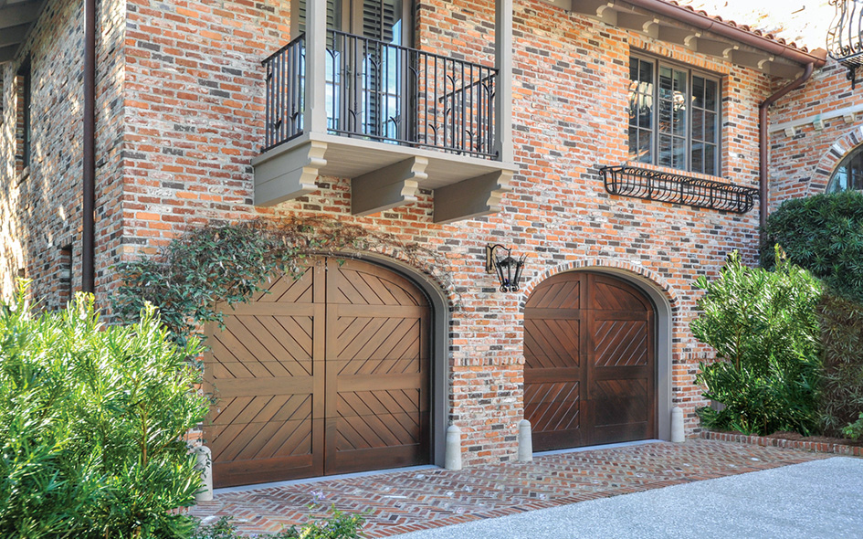 Two Signature Carriage wooden garage doors with chevron and vertical patterns grace a brick house, set beneath a small balcony with black railing and surrounded by lush green shrubs.