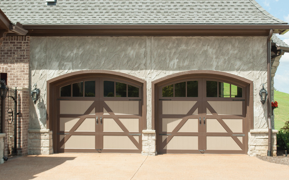 Two adjacent Signature Carriage arched garage doors with brown trim and beige panels, set in a stone and stucco exterior wall beneath a shingled roof. The concrete driveway is well-lit by daylight.