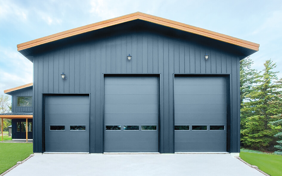 A modern dark blue building with three large insulated garage doors from the Thermacore Collection, each topped with an exterior light, sits on a concrete driveway with green grass and trees in the background.