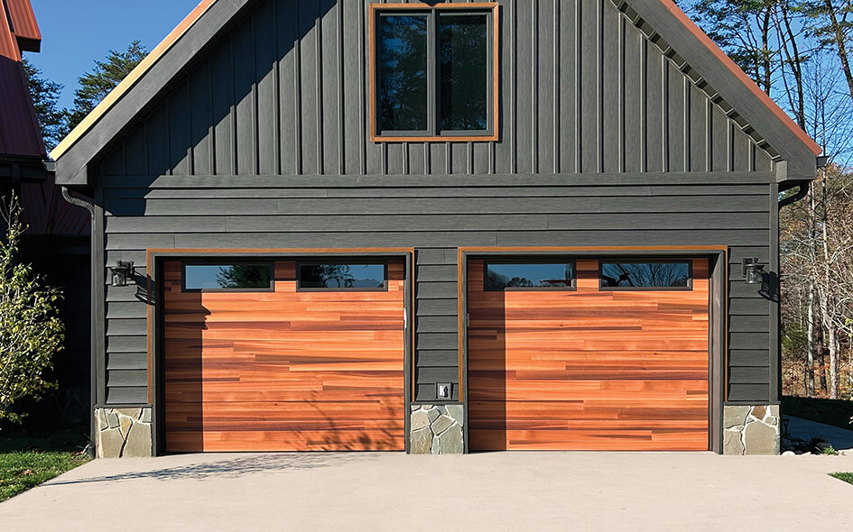 A modern house with dark vertical siding and two Thermacore Collection garage doors, each featuring horizontal panels and rectangular windows at the top. There are stone accents at the base and a double window above the garages.