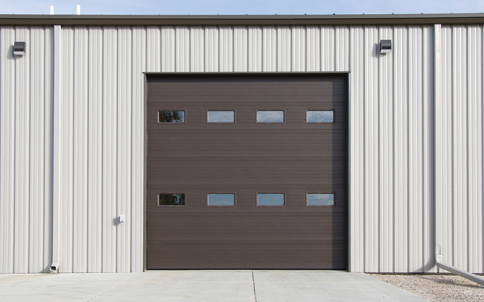Large brown Model 850 industrial garage door with four small rectangular windows is set in a light gray metal building with vertical siding. Concrete pavement leads up to the Advanced Performance Door, and two wall-mounted lights are visible above.