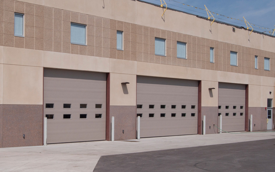 A beige commercial building with three large Thermacore Model 850 garage doors, each featuring four rows of windows, sits under a clear blue sky. The surrounding area is paved with concrete and asphalt.